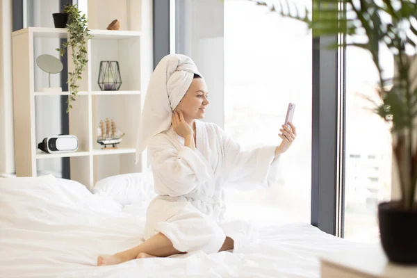 stock image Charming serene woman in after-bath wear holding cell phone while relaxing on soft bed in bright studio apartment. Smiling adult female taking self-portrait in home interior using mobile camera.