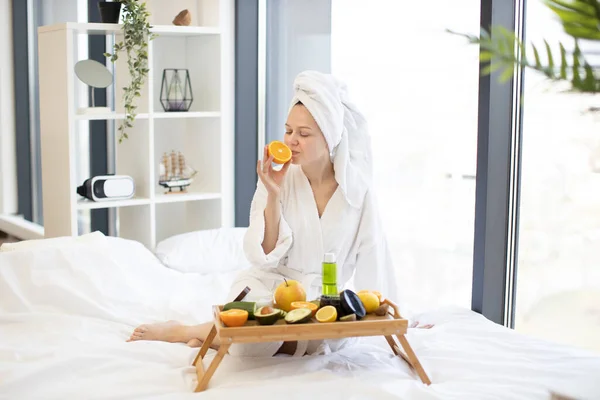 Beautiful young lady in after-bath wear looking at citrus half while sitting on bed with tray table full of skincare products at home. Lovely woman intending to moisturize skin with organic remedy.