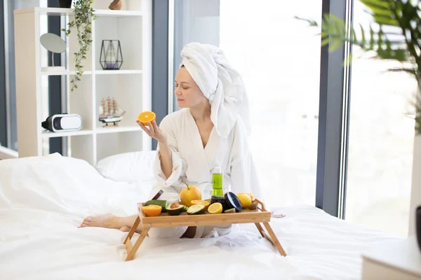 Beautiful young lady in after-bath wear looking at citrus half while sitting on bed with tray table full of skincare products at home. Lovely woman intending to moisturize skin with organic remedy.
