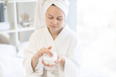 Bornoz ve yumuşak havluyla çekici sakin bir kadın kozmetik kavanozundan yüz kremi alırken kendini spa prosedürleriyle şımartıyor. Sakin kadın, sağlıklı cilt bakımı alışkanlıkları geliştiriyor..