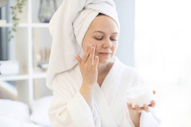 Banyodan sonra giyinen güzel bir bayan stüdyo dairesinin parlak odasında otururken yüz yanağına kremi sürüyor. Alımlı sakin kadın günlük rutin vücut nemlendiricisini takip ediyor..