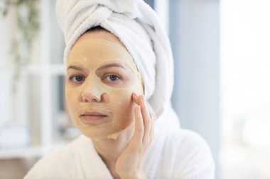 Beyaz bornoz ve havlu giymiş beyaz kadın evde boş vakit geçirirken uygulamalı kağıt maskeyle yüzüne dokunuyor. Yatak odasının iç kısımlarında güzellik hizmetlerinden yararlanan genç kişi.