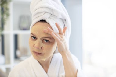 Beyaz bornoz ve havlu giymiş beyaz kadın evde boş vakit geçirirken uygulamalı kağıt maskeyle yüzüne dokunuyor. Yatak odasının iç kısımlarında güzellik hizmetlerinden yararlanan genç kişi.