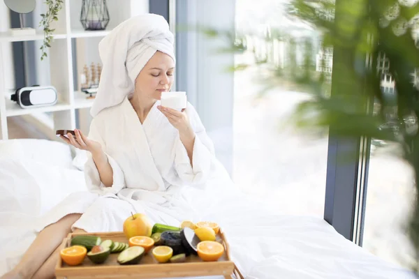 寝室のパノラマの窓から太陽の光を楽しみながら 光の保湿剤の新鮮な香りを感じる風呂の後の魅力的な健康的な女性 美容療法の主な役割を果たす果物のスライス — ストック写真