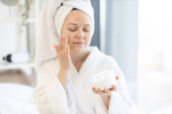 Beautiful young female in after-bath wear applying skincare cream on face cheek while sitting in bright room of studio apartment. Charming calm woman following daily routine of body moisturising.