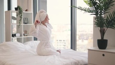 Beyaz sabahlık giymiş gülümseyen yetişkin kadın stüdyo dairesinin parlak odasında rahat yatak örtüleri üzerinde dinlenirken bacaklarını esnetiyor. Neşeli ev hanımı evde eğlenerek hayat memnuniyetini arttırıyor..