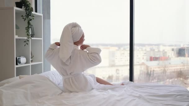 穿着浴巾和白袍的年轻女士坐在通风卧室的全景窗前 手拉手的背景图 快乐的女人在新的一天开始前缓解紧张 — 图库视频影像
