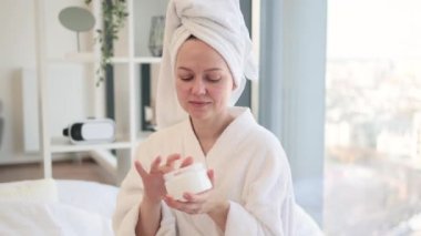 Beyaz banyo havlusu ve bornozuyla parlak arka planda duş aldıktan sonra yüz kremine yapışmış çekici beyaz kadın görüntüsü. Sağlıklı bir kadın cildini sulandırıyor..