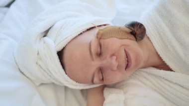 Kafasında banyo havlusu olan güzel bir kadının, yumuşak bir battaniyenin üzerinde yatarken yüzünde sevimli bir salyangoz tutuşu. Huzurlu genç yetişkin cilt üzerinde iyileştirici sümük kullanarak kozmetoloji tedavisi görüyor..