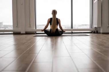 Dar spor elbiseli, açık renk saçlı genç bir kadının spor salonunun penceresinin önündeki yoga minderinde poz verişi. Rahat yogini derin meditasyon için Padmasana egzersizi yapıyor..
