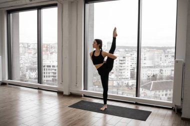 Beyaz geniş stüdyoda trivikramasana pozu veren beyaz saçlı, gelişmiş yoga pratiği yapan bir kadın. Profesyonel yogi rahat spor kıyafetleri giyer, tek bacağıyla dengede durur ve Asana yapar..