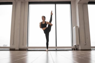 Beyaz geniş stüdyoda trivikramasana pozu veren beyaz saçlı, gelişmiş yoga pratiği yapan bir kadın. Profesyonel yogi rahat spor kıyafetleri giyer, tek bacağıyla dengede durur ve Asana yapar..