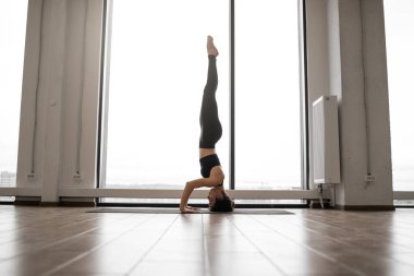 Yoga yaparken konsantre olmuş güçlü bir kadın. Salamba sirsasana 'da kafasının üstünde parlak ışıklı odada poz veriyor. Spor kıyafetleri içinde genç bir kadın vücudunu güçlendiriyor ve beynini sakinleştiriyor..