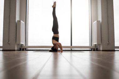 Formda ve ince bayan, siyah dar taytlarla yoga egzersizleri yapıyor ve Fransız pencerelerinin önünde üstte. Parlak emtia stüdyosunda amuda kalkan esmer bir sporcu. Kapalı alanda eğitim kavramı.