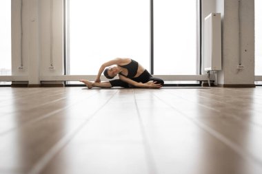 Beyaz tenli bir kadın geniş bir odada yoga minderinde otururken bacaklarını esnetiyor. Spor kıyafetli genç bayan vücut esnetme egzersizleri yapıyor..