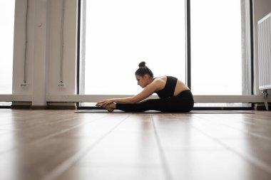 Eğri büğrü genç esmerin panoramik pencerelerin yanındaki yoga minderinde esneme egzersizi yapışının yan görüntüsü. Siyah spor giysiler giyen beyaz bir kadın günlük antrenman rutinini takip ediyor..