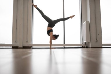 Genç bir kadın panoramik pencereden şehir manzaralı spor salonunda tek başına antrenman yapıyor. Siyah pantolonlu atletik bir kadın ve yoga minderinin üzerinde geniş bacakları açık bir şekilde amuda kalkma pozu..