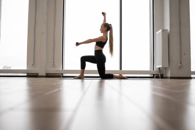 Bir dizi yoga minderinde duran güçlü beyaz kadın ve kol egzersizleri için dambıllar kullanıyor. Siyah giyimli iyi eğitimli bir bayan şehir manzaralı panoramik pencerelerin yanında egzersiz yapıyor..
