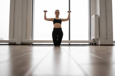 Yoga minderinde dizlerinin üzerinde duran ve kollarını dambıllarla çalıştıran genç bayanın ön görüntüsü. Kafkas fitness kadını rahat aktif kıyafetler giyer stüdyoda spor egzersizleri yapar..