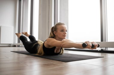 Aktif Kafkasyalı kadın yoga minderine doğru eğiliyor ve elinde dambıllarla geriye doğru eğiliyor. Spor kıyafetleri içinde güçlü genç bir sarışın. Havalandırma odasında yoğun idman yapıyor..