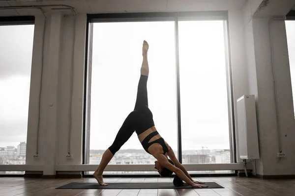 Young caucasian woman in active wear holding Standing Split Pose on yoga mat against background of panoramic window. Urdhva Prasarita Eka Padasana position challenging body focus and balance.