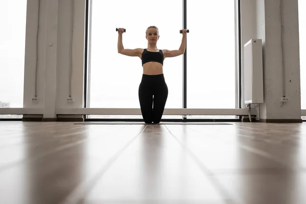 站在瑜伽垫上跪着 用哑铃训练手臂的年轻女子的正面形象 身着舒适活跃衣服的白人健身妇女在演播室里进行体育锻炼 — 图库照片