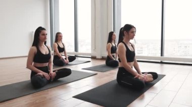 Parlak stüdyoda panoramik pencereli, bacak bacak bacak üstüne atmış bir şekilde meditasyon yapan güzel bayanların yan görüntüsü. Spor yapan yetişkinler yoga dersinde paspaslar üzerinde Padmasana egzersizi yapıyorlar..