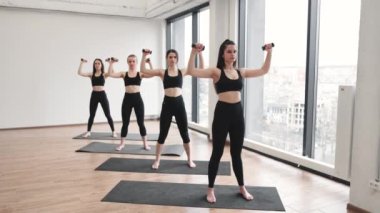 Aktif giyinen bir grup genç kadın dambılları omuzlarının üzerinden tutarak ayakta poz veriyorlar. Aktif sıska bayanlar spor salonunda ağırlıklarla yoga yapıyor..