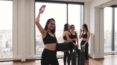 Kolunun altında siyah paspası olan beyaz bir kadın spor şişesini uzatırken yoldaşları panoramik pencerenin yanında gevezelik ediyor. Atletik kadınlar stüdyodaki grup yoga dersine ara veriyorlar..