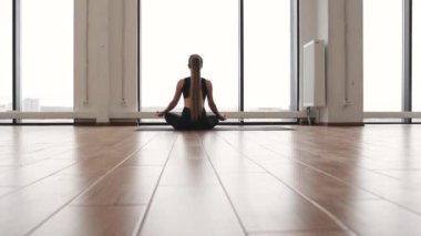 Dar spor elbiseli, açık renk saçlı genç bir kadının spor salonunun penceresinin önündeki yoga minderinde poz verişi. Rahat yogini derin meditasyon için Padmasana egzersizi yapıyor..