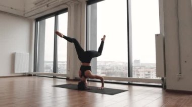 Genç bir kadın panoramik pencereden şehir manzaralı spor salonunda tek başına antrenman yapıyor. Siyah pantolonlu atletik bir kadın ve yoga minderinin üzerinde geniş bacakları açık bir şekilde amuda kalkma pozu..
