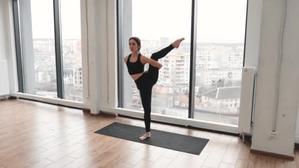 白种人黑头发的女人站在雪白宽敞的画室里练习高级瑜伽 职业瑜伽选手穿着舒适的运动服 一条腿上保持平衡 — 图库视频影像