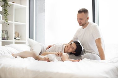 Günlük giysiler içinde yakışıklı bir erkek. Parlak yatak odasındaki yumuşak yorganın üzerine uzanmış küçük bir ailenin gülümseyen dişilerine şefkatle bakıyor. Evli ve çocuklu mutlu çift içeride eğleniyor..