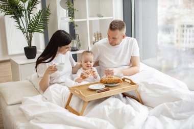 Küçük kızının kruvasan tatmasını izlerken güzel annenin beyaz battaniyeye konmuş yatak tepsisinden içmesinin keyfini çıkarmasına şaşırdım. Genç yetişkinler ve tatlı bebek yatakta hafif yiyecekler yiyorlar..