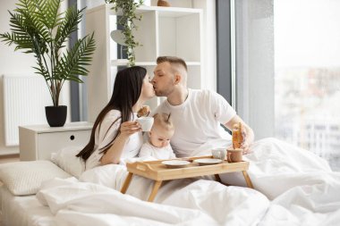 Çekici evli çift birbirlerini öperken küçük kız stüdyo odasında battaniyenin altında ebeveynlerin arasında oturuyor. Üç kişilik neşeli bir aile pazar sabahını yatakta birlikte geçirir..