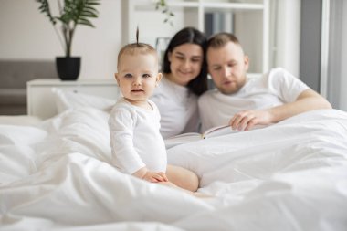 Beyaz bebek kıyafetleri içindeki neşeli küçük çocuk yatakta dinlenirken neşeli yetişkin insanlar aydınlık yatak odasında battaniyenin altında kalıyorlar. Sevgi dolu anne ve baba şiir kitabından çocuk tekerlemesi anlatıyor..