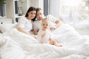 Beyaz bebek kıyafetleri içindeki neşeli küçük çocuk yatakta dinlenirken neşeli yetişkin insanlar aydınlık yatak odasında battaniyenin altında kalıyorlar. Sevgi dolu anne ve baba şiir kitabından çocuk tekerlemesi anlatıyor..