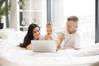 Her gün giyilecek kıyafetleri olan neşeli evli çift hafta sonu boyunca tatlı bir kızla yatakta dinlenirken modern dizüstü bilgisayar kullanıyor. Genç ebeveynler internette sörf yaparken küçük çocuklar sağlıklı atıştırmalıklar yiyor..