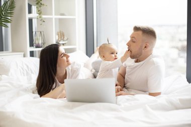 Sevimli tatlı çocuk, yakışıklı babaya günlük kıyafetle meyve jölesiyle davranırken mutlu anne de stüdyo dairesindeki modern dizüstü bilgisayarını çalıştırıyor. Bakıcı ebeveynler evde bebekle dinleniyor..
