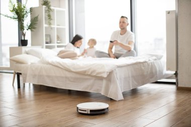Esmer kadın bebek kızla yatakta oynarken genç adam stüdyo odasındaki uzaktan kumandayla robot elektrik süpürgesini açıyor. Modern aile hafta sonları evde temizlik yaparak dinleniyor..