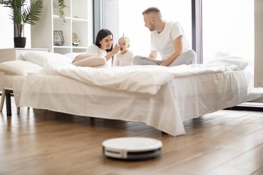 Rahat eşler küçük çocukları kaşıkla beslerken akıllı robot süpürgesi de stüdyo dairesindeki plastik zeminden tozu temizliyor. Elektronik temizlik teknolojisi kullanan bebekli modern aileler..