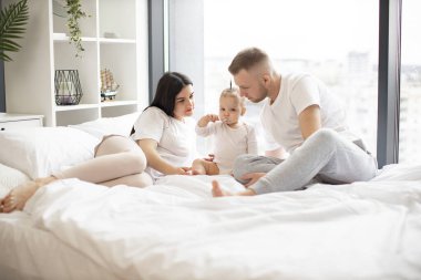 Güneşli bir günde anne ve babasıyla gündelik kıyafetleriyle yatakta otururken kaşıkla yemek yiyen sevimli bir bebek. Evde kalırken küçük kızına özen gösteren ebeveynler..