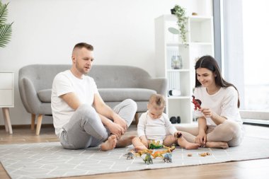 Genç, evli bir çift oturma odasında bacak bacak üstüne atarak oturuyor ve halının üzerinde küçük bir kızla dinozor oyunu oynuyorlar. Üç kişilik sevgi dolu aile evde birlikte vakit geçiriyorlar..