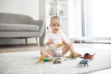 Oturma odasındaki yumuşak halıda dinlenirken rengarenk hayvanlarla poz veren günlük bebek kıyafetleri içindeki şirin çocuk portresi. Evde dinazor oyuncakları oynayan çıplak ayaklı mutlu kız..