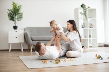 Neşeli eşler beyaz günlük kıyafetler giyip yumuşak halılı, yumuşak, şirin bir bebek kızı tutuyorlar. Dinozor oyuncakları ve tahta bloklar etrafa saçılmış. Genç bir ailenin eğlencesi ve aşkı.