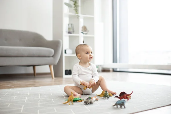 Delightful baby girl in white bodysuit sitting on floor carpet and holding toy dinosaur in hands. Adorable infant child with funny ponytail staying calm and happy on sunny day at home.