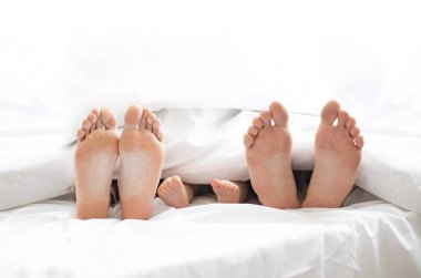 Kapalı alanda rahat bir yatakta, beyaz yumuşak bir battaniyenin altında gizlenmiş üç çift çıplak ayak görüntüsü. Mutlu anne, baba ve bebek evde güneşli bir sabahta birlikte dinleniyorlar..