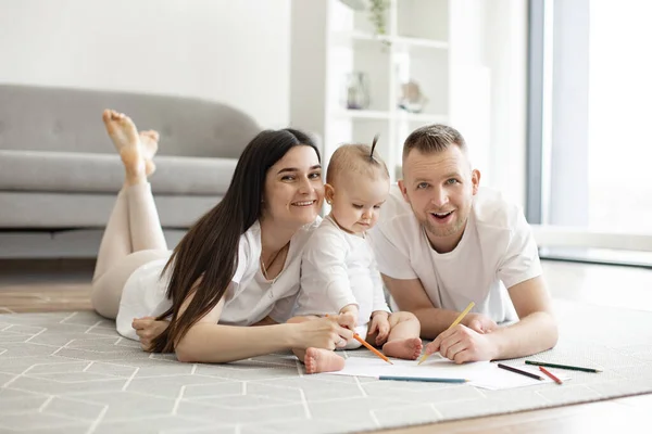 stock image Smiling family of three in casual attire posing with drawing pencils while relaxing on wooden floor in studio apartment. Funny caucasian spouses developing babys imagination using art and colors.