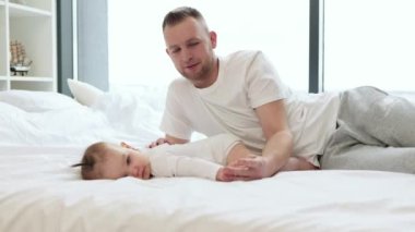 Küçük çocuğu karnında tutan yakışıklı sakallı adamın portresi. Nazik bir baba ve beyaz elbiseli güzel bir kız bebek yumuşak bir yatağa uzanıp kameraya bakıyor. Ebeveyn sevgisi ve bakımı kavramı.