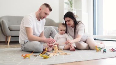 Rahat bir apartmanın zemininde oturan ve küçük tatlı kızıyla oynamak için tahta bloklar kullanan, sıradan giyimli beyaz ebeveynler. Evde yeni yürüyen çocuk için eğitici bir oyun.